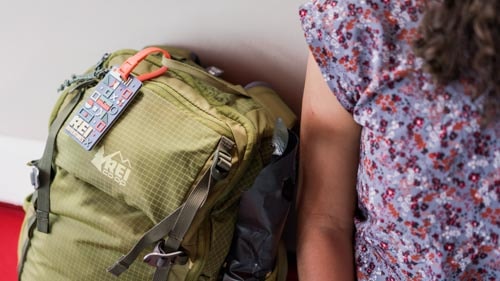 A green backpack with an R E I luggage tag sits next to a traveler
