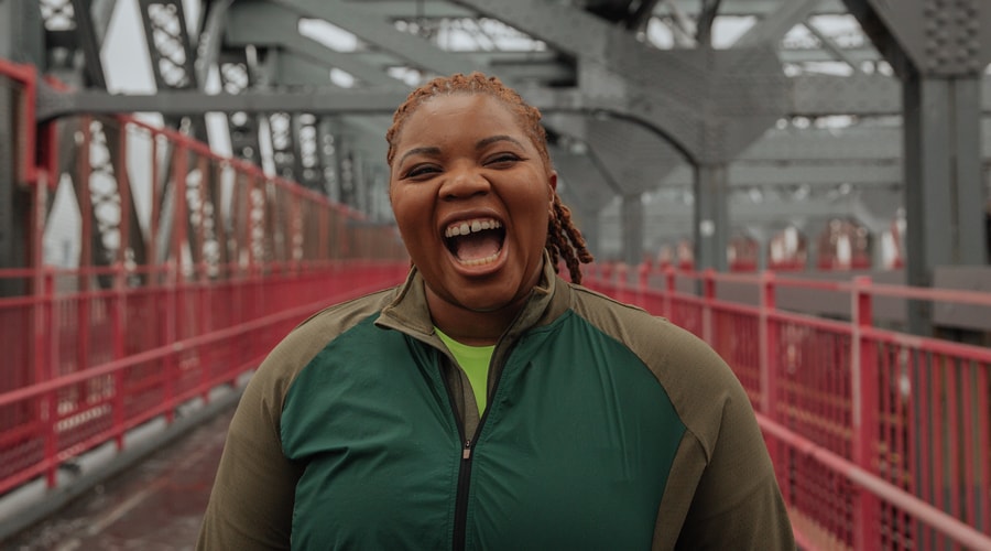 A woman wearing a fitted technical long-sleeve shirt laughs while standing on a bridge's pedestrian trail.