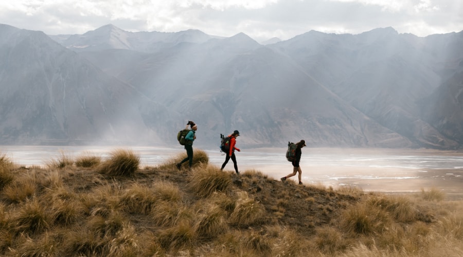 Three hikers wearing packs traverse a gentle slope against a dramatic backdrop of water, layered triangular peaks, and sunbeams breaking through clouds.