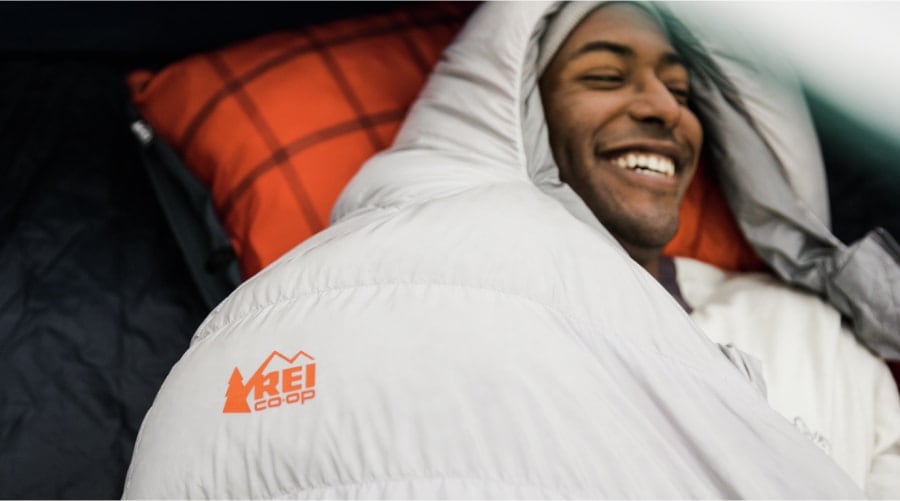 A man smiles while snuggled up in an REI Co-op brand mummy-style sleeping bag, on top of a colorful camp pillow.