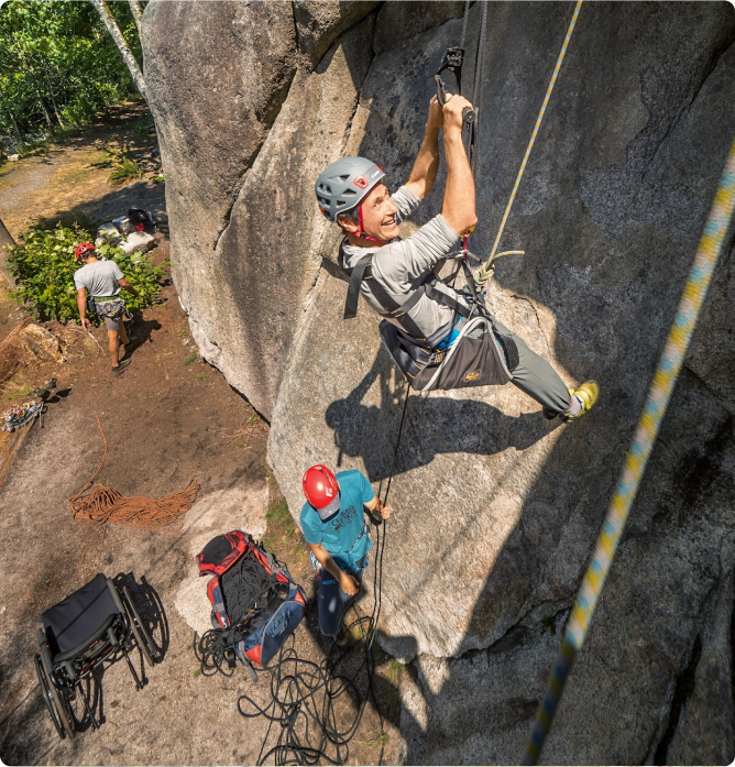 A rock climber strapped into harness smiles on the side of a rock.
