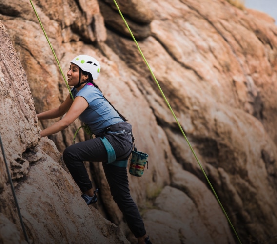 A climber scales the face of a rocky cliff. They are wearing a helmet and are strapped into a climbing harness.