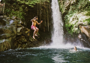 a man jumps off of a rock into a pool of water at the foot of a waterfall