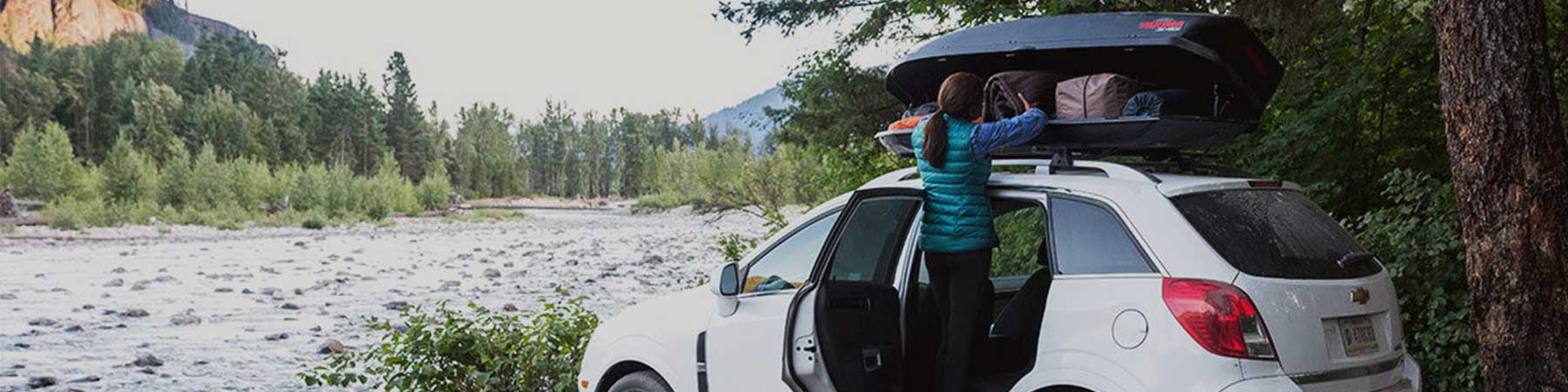A camper offloads gear from her car's roof-top box next to a river