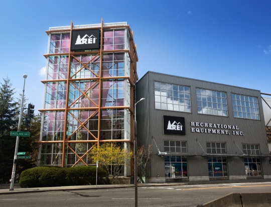 The front of an R E I store against a backdrop of trees, mountains, and clear sky