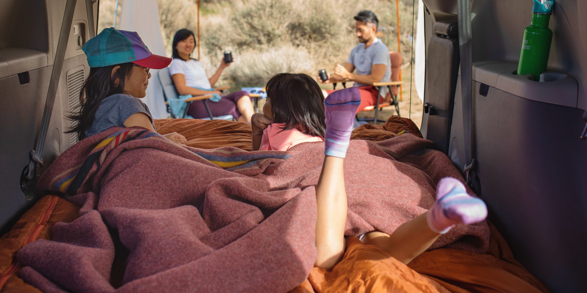 Two kids sprawl in the back of a camper van while two adults sip and supervise them from beneath a sun canopy
