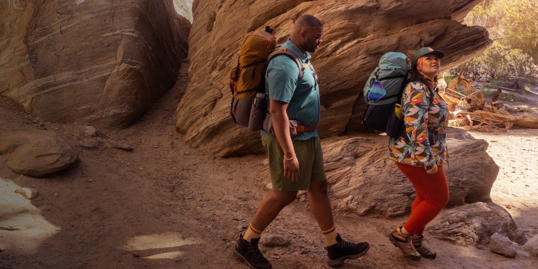 Two backpackers hiking through a sunlit sandstone canyon.