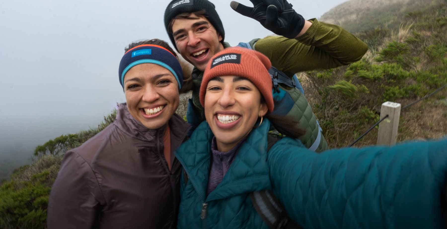Three people bundled up in beanies and jackets, smiling and taking a selfie on a hiking trail.