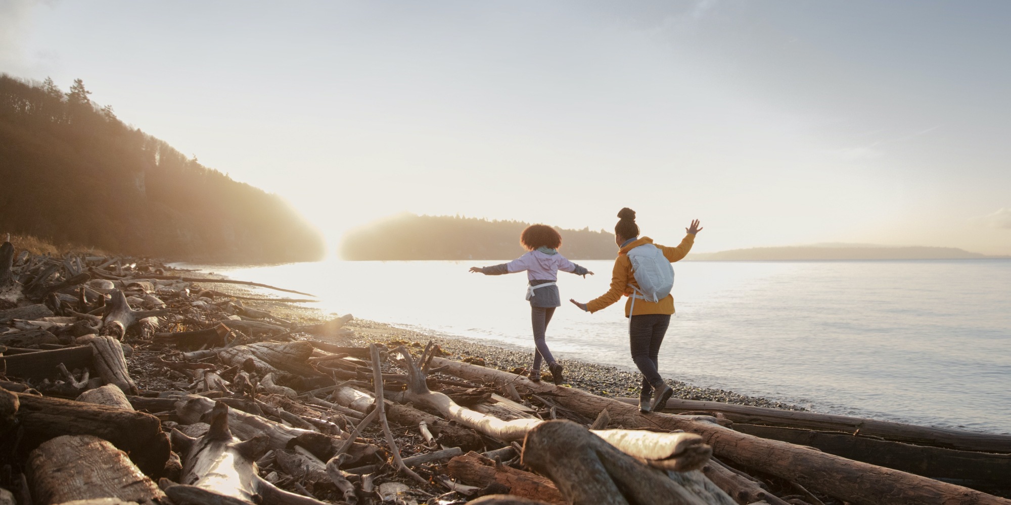 Two girls walk on driftwood on a pebble-strewn beach near the water.