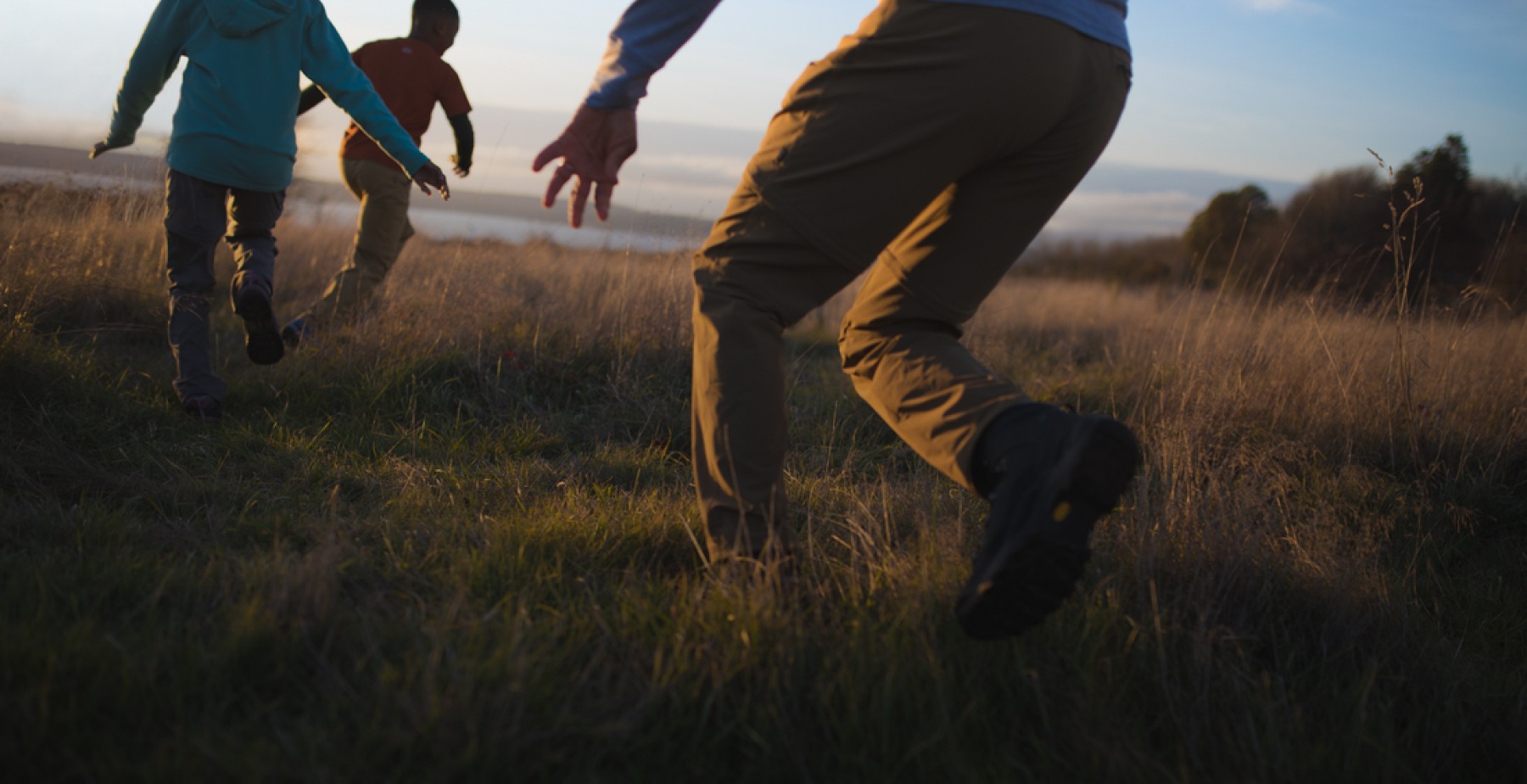 Children running in tall grass at magic hour, with water and trees in the background.