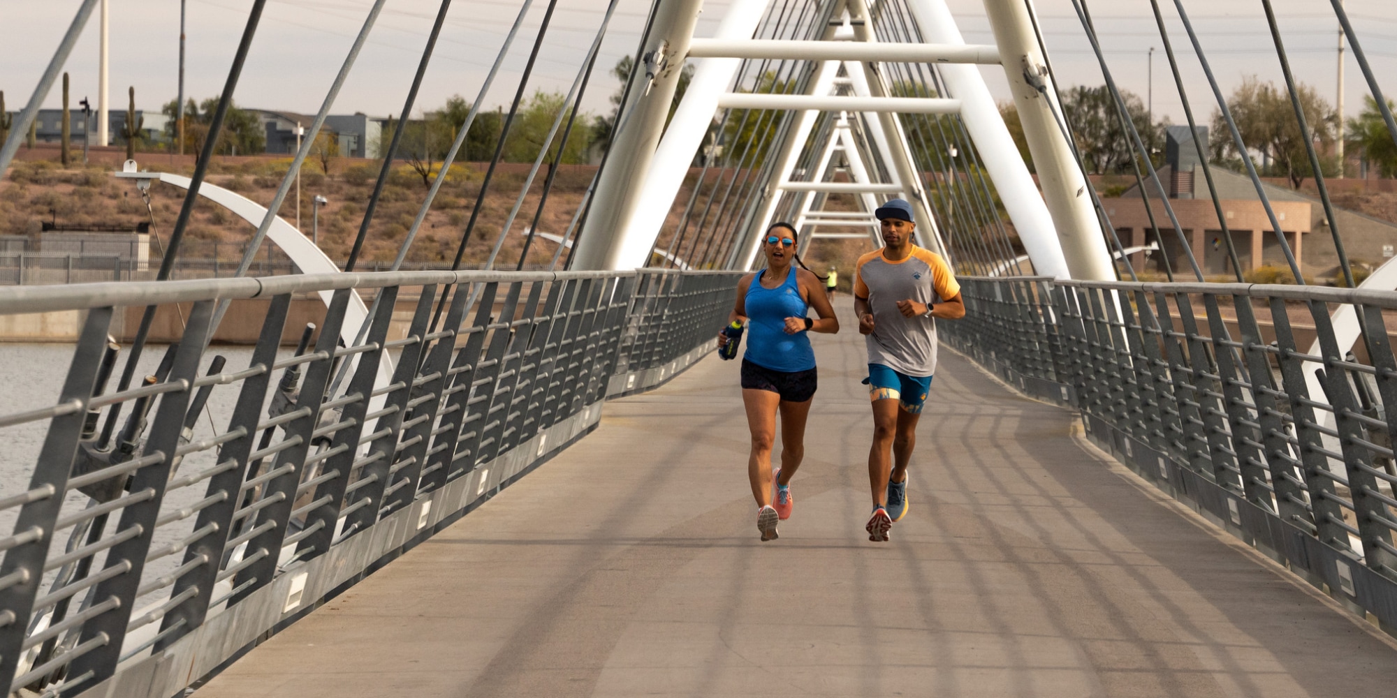 Two runners cross a bridge over a river on a sunny day.