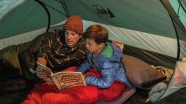 Under the glow of a camp lamp, father and son share a book in their tent.