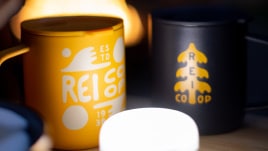 Two REI Co-op branded mugs sit on a table next to a fire.
