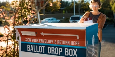 A white woman drops her mail-in ballot into an outdoor ballot drop box.