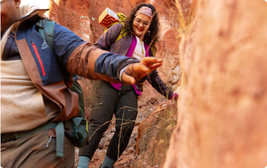 Two hikers navigate a narrow, sun-dappled red stone canyon.