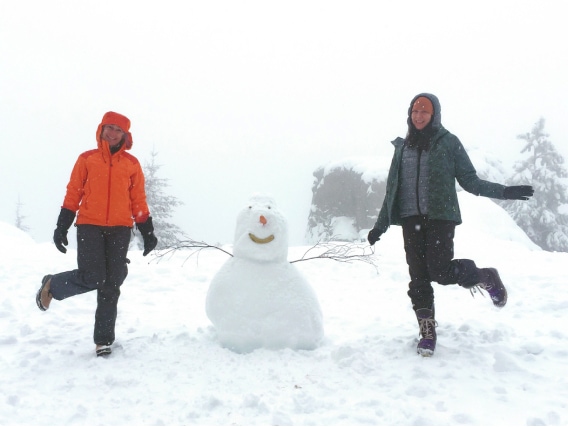 Two people pose with a smiling snowman in a snow-covered field bordered by coniferous trees.