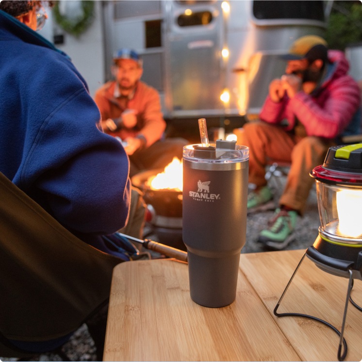 A happy camper in a puffy head-to-toe onesie stands with his arms wide next to a campfire, while two other seated campers smile up at him.
