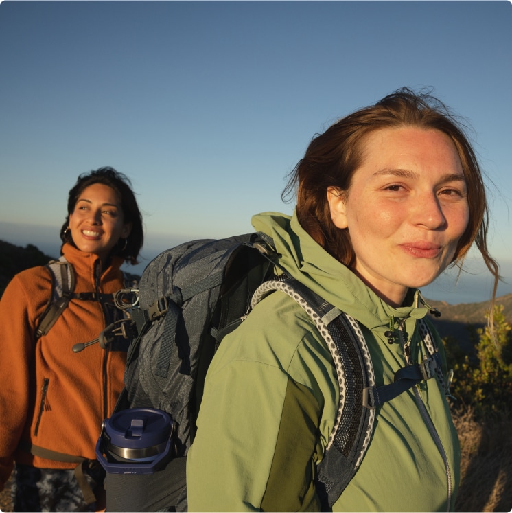 Three smiling hikers in mid-weight jackets and long pants on a dirt trail bordered by scrub on a grey day.