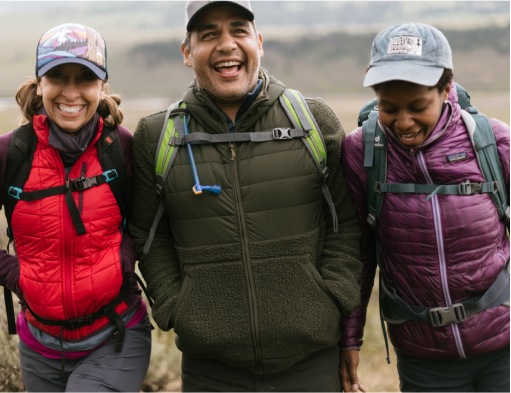 Three hikers stand side by side laughing while wearing hats, puffers and backpacks on a chilly overcast day.