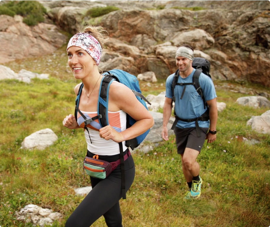 Two young adults in backpacks and lightweight outdoor apparel hike on a grassy slope studded with large rocks