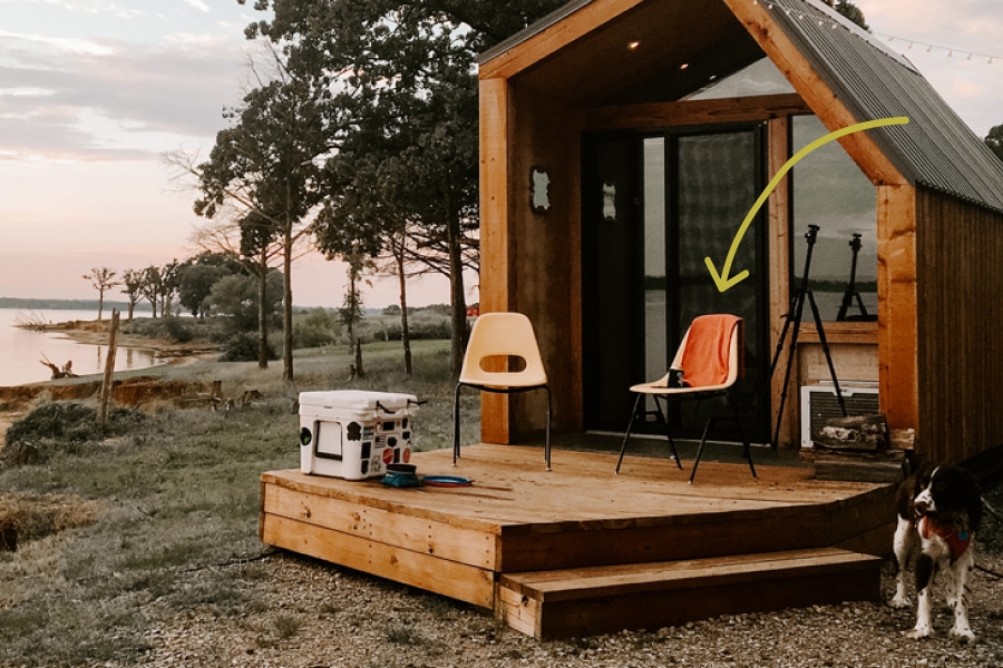 Two chairs sit invitingly on the deck of a rustic-modern tiny cabin while a dog waits nearby, mouth open, tongue out.