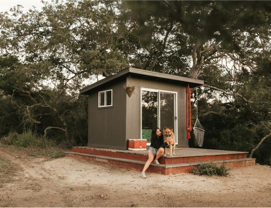A person hugs their dog on the front deck of a tiny house in front of large trees