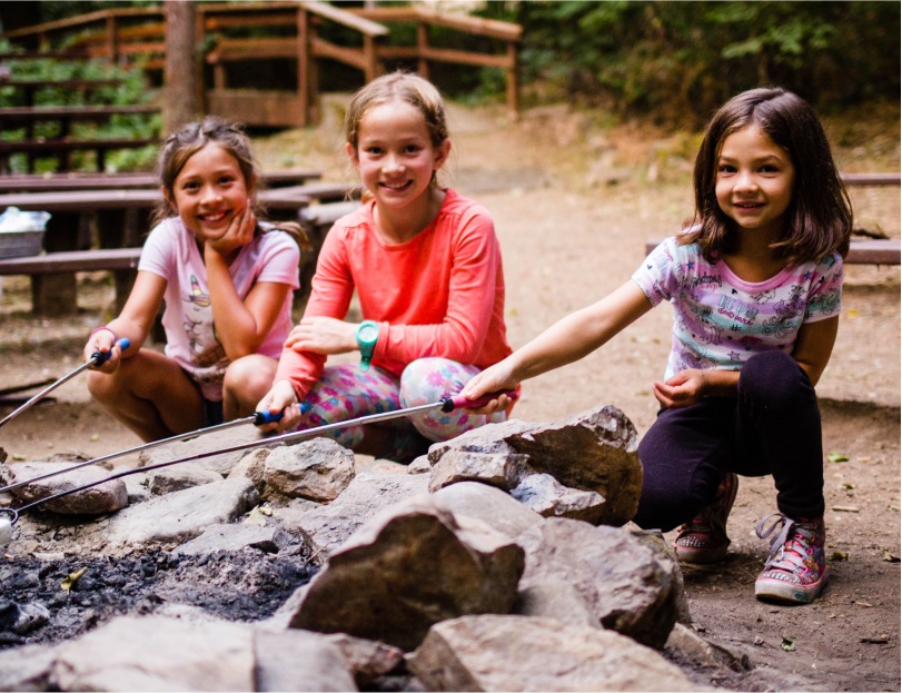 Three smiling kids roasting marshmellows over a campfire.