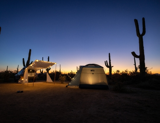 A tent and tailgate awning glow with string lights at a desert campsite at dusk