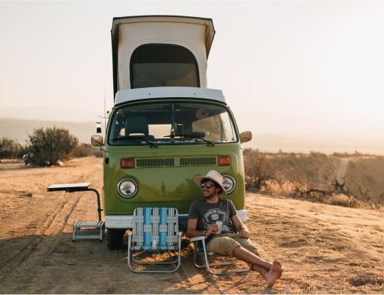 A camper lounges in a camp chair in front of their van on a remote dirt road.
