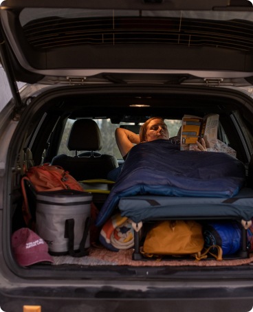 A car camper rests inside their sleeping bag reading.