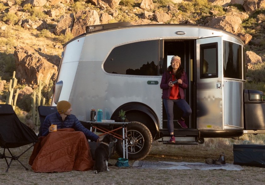 The R E I Co-op Special Edition Basecamp Trailer. A group of campers drink coffee outside their Airstream trailer.
