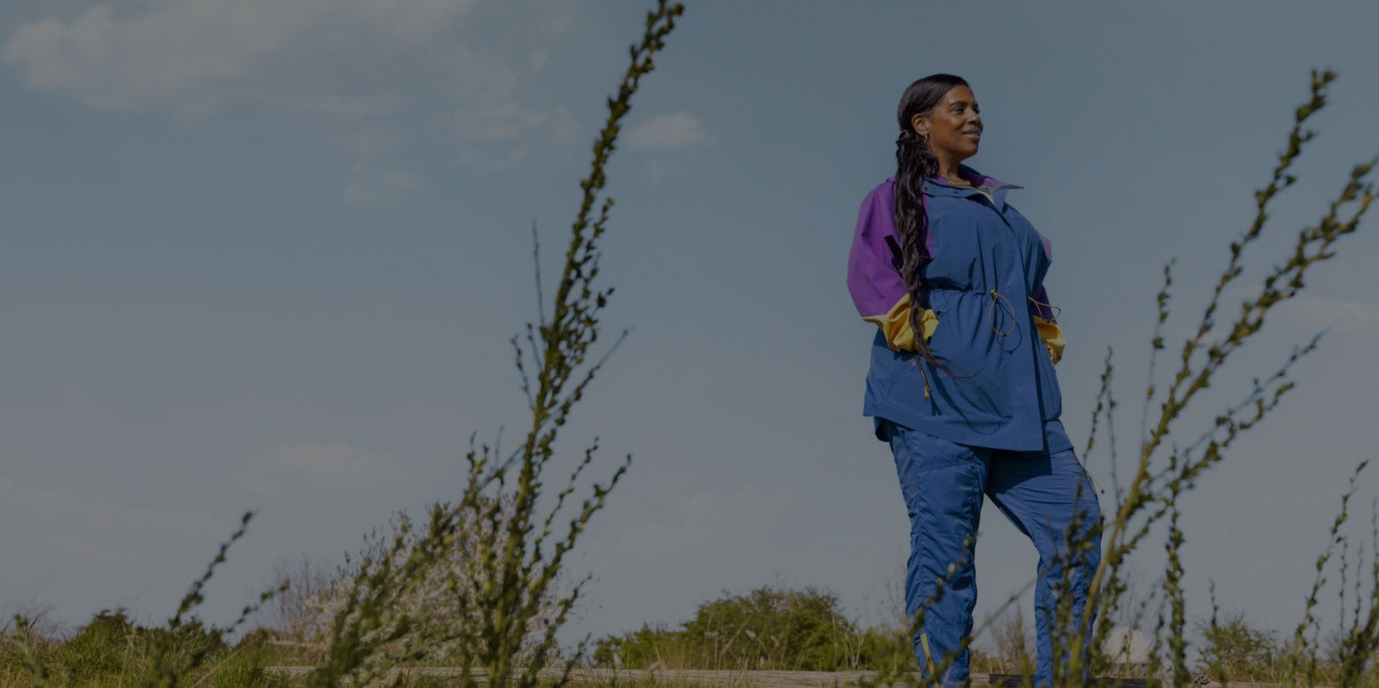 A person smiling and looking out in a grassy field, while wearing the Outdoor Afro Inc. plus R E I Co-op collection.