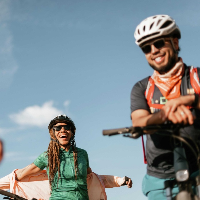 Two mountain bikers pause for a laugh under blue skies and puffy clouds.