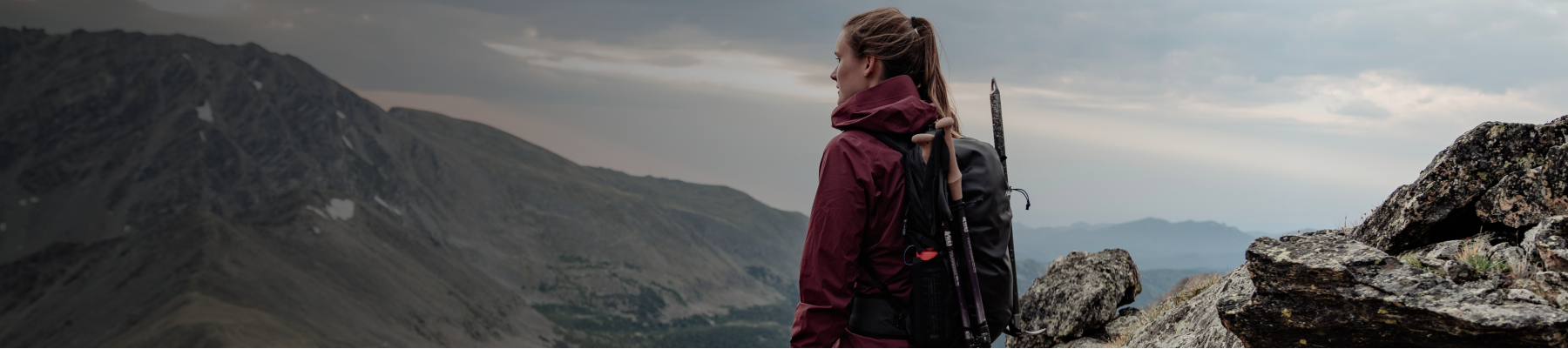 A woman wearing midweight technical outdoor apparel and a pack with attached poles looks out over a muted mountain landscape with a lake, snow patches, and marbled sky