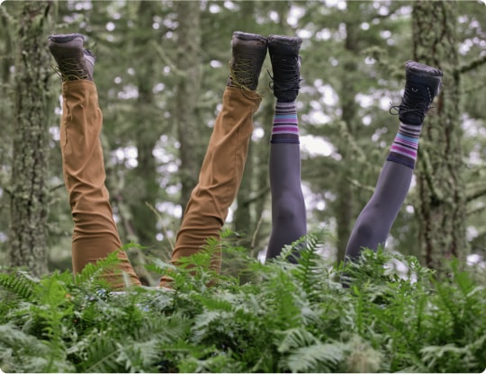 Against a forest backdrop, with bright green ferns in the foreground, two pairs of legs dressed in midweight outdoor attire and hiking boots stick up in the air, implying two people behind the ferns doing headstands