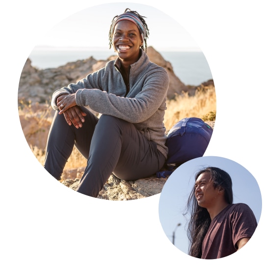 A smiling woman wearing midweight outdoor athletic attire sits on a rock amidst a landscape of rock, scrub brush, and clear blue sky