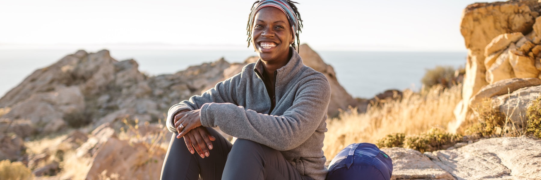 A smiling woman of color wearing midweight outdoor gear sits on a rock against a desert-like backdrop of scrub, rock outcroppings, calm water, and clear sky.