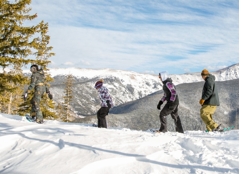 Snowshoers make their way up a trail near the top of a mountain, against a partly cloudy sky.