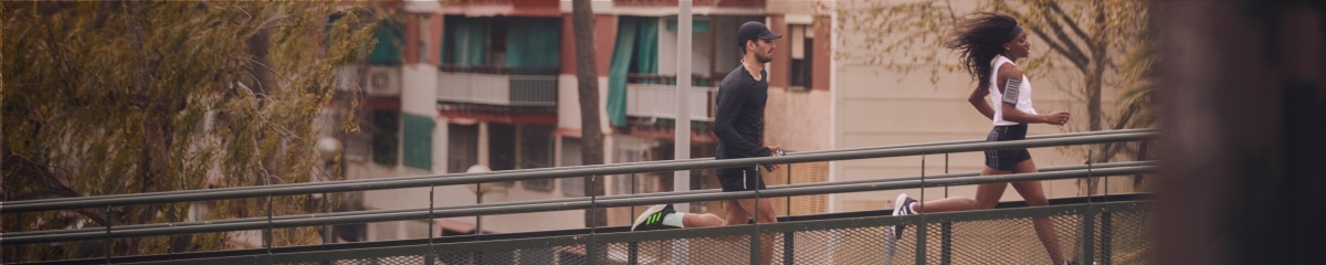 Two runners jog across a pedestrian footbridge in an urban park.