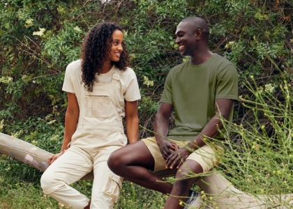 Two people in Outdoor Voices, smiling and sitting on a log, surrounded by greenery.