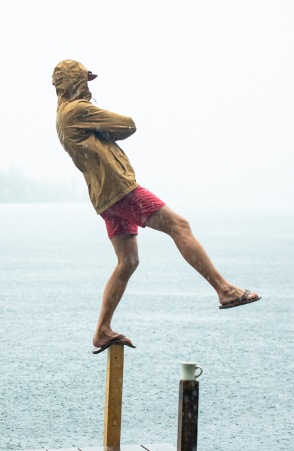 In the rain, a person in a swimsuit and raincoat is balancing on one foot on a pole overlooking the water.