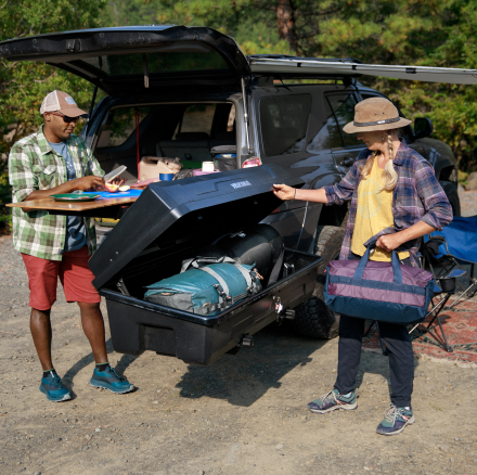 A person carrying multiple bags unloads a duffel bag from a Yakima hitch-mounted cargo box filled with gear.