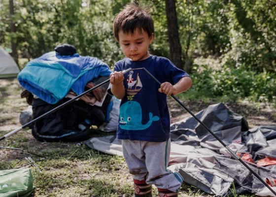 A child standing in a wooded campsite joins two tent poles together.