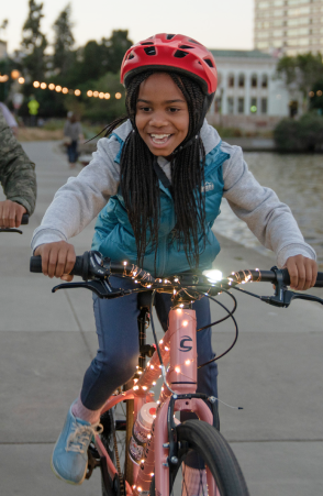A smiling child rides a bike decorated with festive string lights on an urban pathway.