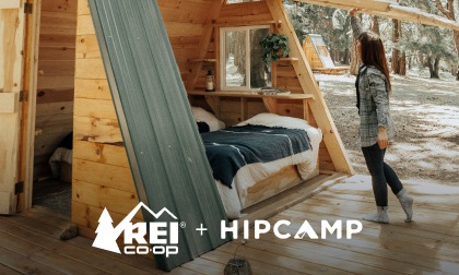 A camper in front of a tiny A frame cabin. Text reads R E I plus Hip camp.