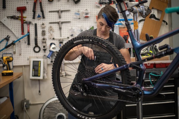 Tools lining the wall behind them, an R E I store bike tech works on a bicycle's derailleur.