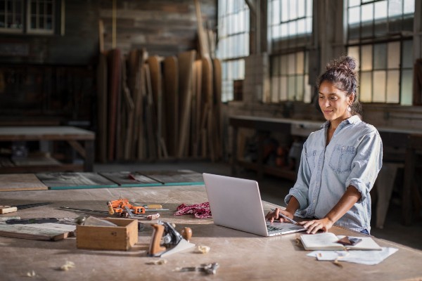 Hand tools strewn on a large workshop table, a product designer consults their laptop as sunlight streams through vintage windows.
