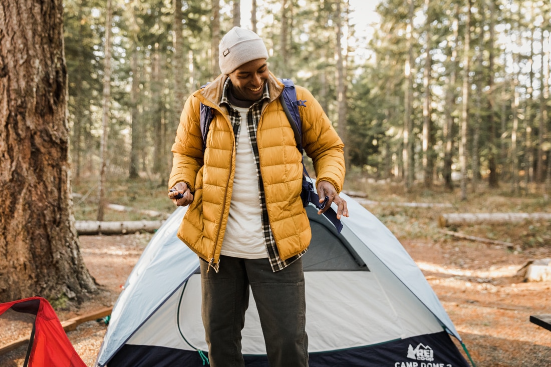 A man wearing an insulated jacket, beanie and backpack stands in front of a tent