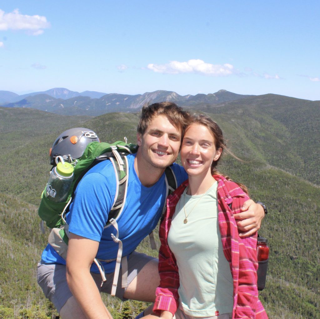 The couple hugs while wearing climbing gear in fronts of a backdrop of green mountains.