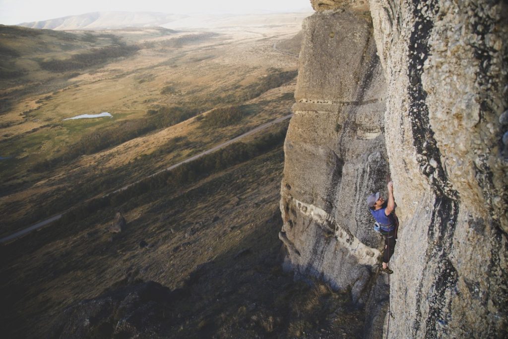 A climber reaches for a hand hold on a cliff face that's bumpy with stone cobbles. A broad valley edged in fog stretches into the background.
